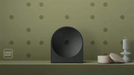 Černý bezdrátový reproduktor s minimalistickým designem a kruhovým čelním panelem, umístěný na olivově zelené polici, pohled zepředu