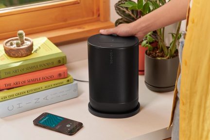 Caixa de som preta cilíndrica sobre mesa branca, mão pousada em cima, livros empilhados, planta em vaso, smartphone com tela acesa