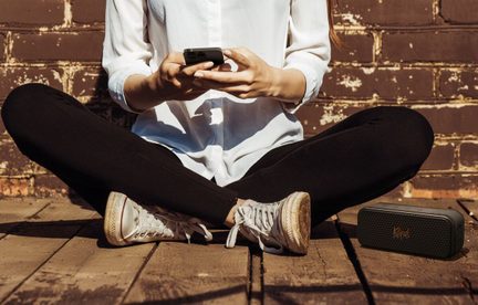Людина сидить, тримаючи телефон, одягнена в чорні штани, біло-сірі кросівки, на дерев'яній підлозі