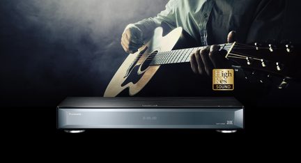Hands playing an acoustic guitar over a Panasonic Blu-ray player with digital display, on a dark background