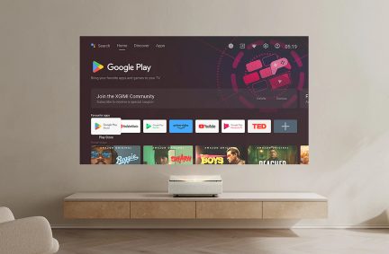 Baltas projektorius ant šviesaus medžio baldų, sieninis ekranas, rodantis „Google Play“ sąsają su programėlių ir filmų piktogramomis.