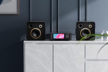 Črni hi-fi komplet, ki vključuje dva kvadratna zvočnika iz mat plastike z zlatimi obročki okoli zvočnikov in osrednji pravokotni predvajalnik z barvnim zaslonom, viden s sprednje strani, vse skupaj postavljeno na svetlo marmorno komodo