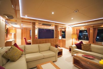 Luksuriøst yachtinteriør med beige sofaer, røde puder, træbordplader, indbyggede TV-skærme og integreret loftbelysning i træpaneler