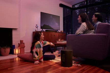 Stue, tre personer foran et tændt tv, sort cylindrisk subwoofer, lilla sofa i stof, trægulv