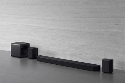 Mattschwarzes Audio-Set aus Kunststoff mit Metallgittern, bestehend aus einer horizontalen Soundbar, einem würfelförmigen Subwoofer mit abgerundeten Ecken und zwei rechteckigen Satellitenlautsprechern, aus leicht erhöhter Dreiviertelperspektive von vorne aufgenommen