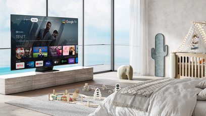 TCL 65C831 2022-fjernsyn, der viser Google TV-grænsefladen, i en stue med sofa, tæppe, dekorativ kaktus og store vinduer