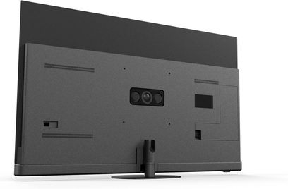 Panasonic TV-65Z90B højopløsnings-tv i antracit-sort, mat plastkabinet, set bagfra i trekvart venstre vinkel med centralt, cirkulært fod og centreret kameramodul med tre linser