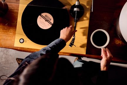 Svetlá drevená vinylová platňa, čierna doska a prenoska, ruka držiaca prenosku a biela šálka kávy, pohľad zhora