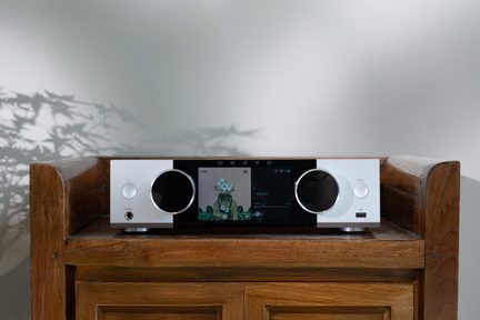Stříbrný síťový Hi-Fi zesilovač Onkyo s velkým barevným centrálním displejem a dvěma velkými kulatými knoflíky na dřevěném stolku