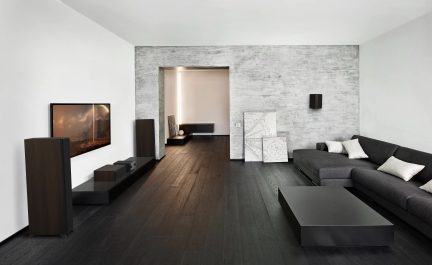 Modern salon, koyu renk ahşap zemin, gri kanepe, siyah dikdörtgen sehpa, dokulu gri duvar, duvar ekranı, hoparlörler