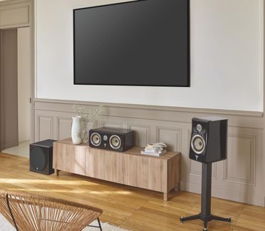 Stue, sort fladskærms-tv monteret på væg, lavt møbel i lyst træ, sort gulvstående højttaler, centerhøjttaler og subwoofer, skråt perspektiv