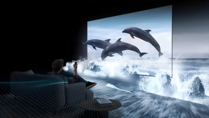 Pantalla plana negra con panel de vidrio ultrafino vista en tres cuartos, imagen 3D de delfines saltando sobre un mar agitado, espectador sentado en un sofá gris en primer plano
