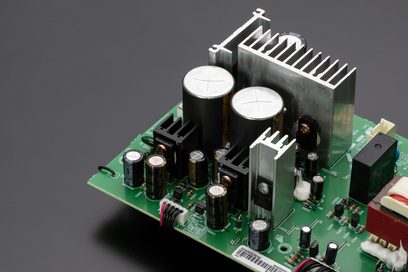 Placa de circuito impresso verde com componentes eletrônicos, dissipadores de alumínio e dois grandes condensadores cilíndricos prateados, vista superior