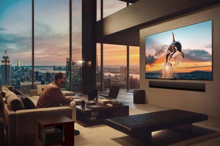 Skråt overblik over en moderne stue med store vinduespartier, der viser et stort sort fladskærms-tv med metalramme monteret på en beige væg over en sort, rektangulær soundbar, en mand siddende i en grå stofsofa overfor et mørkt sofabord i træ, byens skyline i solnedgang i baggrunden