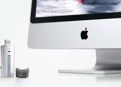 Hvid iMac-computer, sølvfarvet USB-nøgle, sort kabinet med hvidt Apple-logo, skærm der viser et billede af brydende bølger