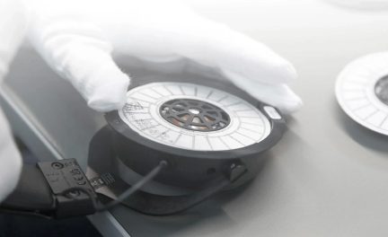 Bela rokavica, ki se dotika črne tuljave z belo etiketo na sivi mizi, priključen črn kabel, pogled od zgoraj