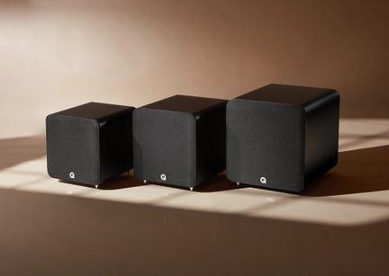 Three black cubic subwoofers, enclosures with a matte finish and dark acoustic fabric fronts with rounded corners, small centered white logo and metal spikes, front view slightly in perspective