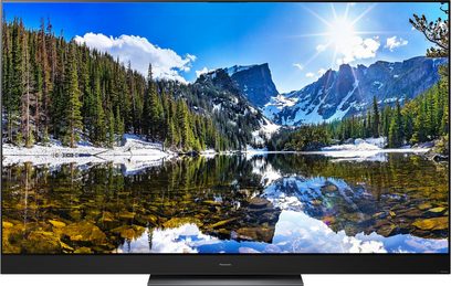 Panasonic TX-65HZ2000E-fladskærm, der viser et bjerglandskab med nåleskov og sø, der spejler den blå himmel og skyer