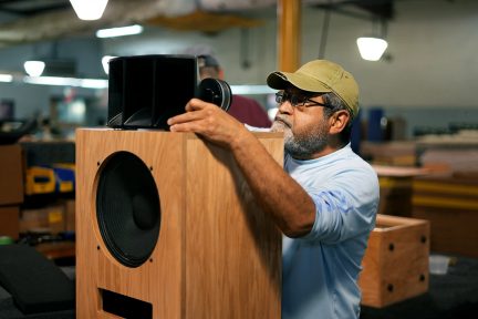Technician installing a black grille on a wooden speaker cabinet with a large visible driver