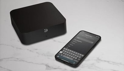 Μαύρο ματ τετράγωνο περίβλημα και smartphone με εφαρμογή ελέγχου Bluesound, άνω όψη σε γκρι μαρμάρινη επιφάνεια