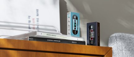 Dva obdĺžnikové audio boxy z tyrkysovo modrého a čierneho plastu, napodobňujúce kazetu so sieťotlačou cievok a bočnými striebornými tlačidlami, zobrazené z trojštvrťového uhla spredu, postavené na stohu CD obalov