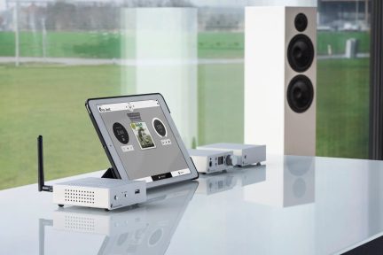Bílý lesklý stůl s nakloněnou deskou zobrazující rozhraní, kompaktní elektronické skříně, vertikální anténa, bílý reproduktor