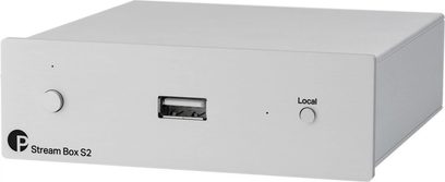 Reproductor de audio en red Stream Box S2 en metal claro, vista frontal en ángulo, botón circular a la izquierda, puerto USB central, botón "Local" a la derecha