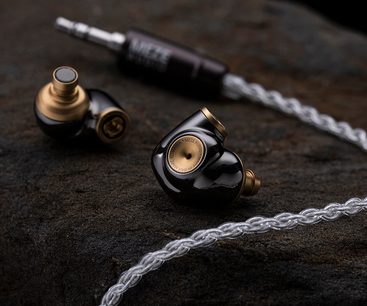 Sorte og guldfarvet Meze Audio ADVAR in-ear hovedtelefoner, med sølvfarvet flettet kabel, på sort tekstureret overflade