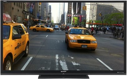 Sharp LC-60LE740E fladskærms-TV, set forfra, stående på et stativ, på en travl gade med gule taxier og fodgængere
