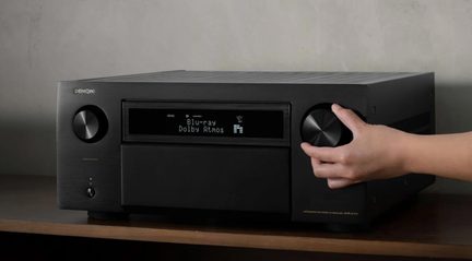 Černý audio-video přijímač z kartáčovaného kovu, pohled z tří čtvrtin zepředu, centrální digitální displej v provozu, dva velké boční otočné knoflíky, z nichž pravý je ovládán rukou