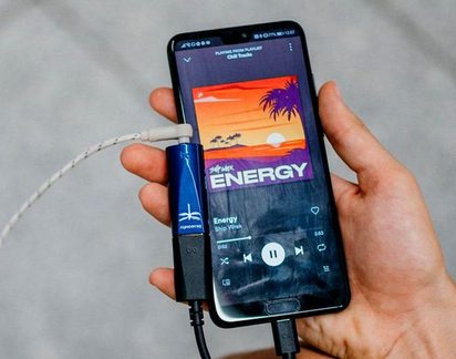 Smartphone nero in mano, schermo di riproduzione musicale, dongle audio blu in metallo e cavo intrecciato collegati, vista a tre quarti