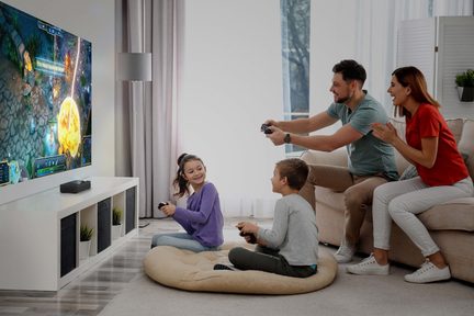 En skærm, der viser et videospil, 2 børn siddende på en pude, 2 voksne på en sofa, alle med controllere i hænderne