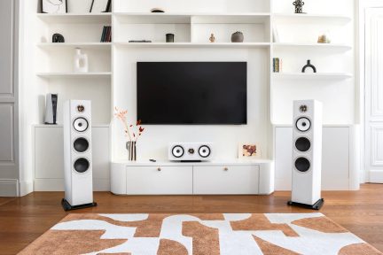 White floorstanding speakers and matching center speaker in front of a white TV stand with a wall-mounted screen in a modern living room