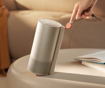 Videoproiettore XGIMI MoGo 3 Pro cilindrico inclinato su tavolo beige, mano che tiene cavo con estremità rossa, libro aperto a destra