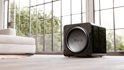 Sort subwoofer i sort, SVS-logo, glat overflade, set fra siden, trægulv, beige sofa, store vinduer