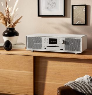 Weißes Sonoro-Hi-Fi-System mit Lautsprechergittern, Knöpfen und Display auf einem Holzregal, schwarze Vasen und Bilderrahmen an der Wand