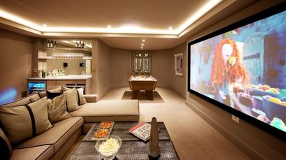 Svetainė su smėlio spalvos kampiniu sofa, stikliniu žurnaliniu staliuku ir projekcijos ekranu, kuriame rodoma raudongalvės garbanotos moters nuotrauka
