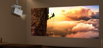 Šedý plastový krychlový miniprojektor s texturovaným povrchem a mikroperforací, s integrovanou horní rukojetí a viditelným předním objektivem, zavěšený na stropě v tříčtvrtečním pohledu zespodu, světelný paprsek směřující doprava