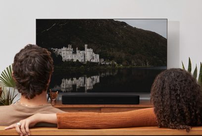 Televisore che mostra un castello sulle rive di un lago che riflette la sua immagine, guardato da due persone di spalle con capelli ricci