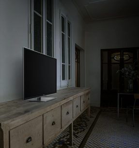 Lyst trækabinet med skuffer foran et vindue, sort tv-skærm placeret ovenpå i et mørkt rum
