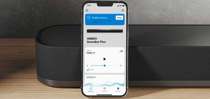 Smartphone s rozsvíceným displejem zobrazující rozhraní aplikace AMBEO Soundbar Plus, položený na černé zvukové liště, světlý dřevěný povrch