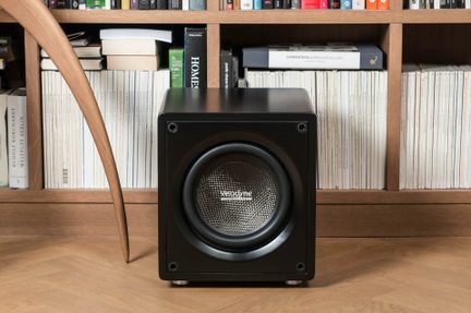 Sort, kompakt Velodyne-subwoofer med firkantet front og centreret højttaler, foran en bogreol i træ