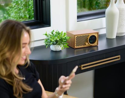 Světlé dřevěné audio zařízení, pohled zepředu, diagonální mřížka, kruhový displej a zlaté tlačítko, na černé konzoli