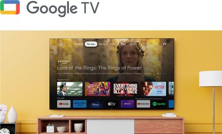 Fladskærms-tv monteret på væggen med Google TV-interface, gul væg, lavt møbel, hvid lampe, vaser, hylder, dekorative genstande
