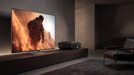 Black home theater projector placed near a television displaying an action scene
