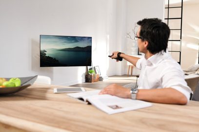 Man zittend aan een bureau, wijzend naar een tv-scherm waarop een zeelandschap met bergen te zien is