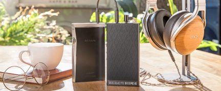 Cuffie audio in legno e metallo su supporto, lettore audio nero, amplificatore nero con texture, tazza bianca, occhiali dorati, taccuino marrone