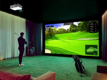 Indendørs rum med storskærm, der viser en golfbane, grøn måtte, ophængt projektor, golftaske, person stående med golfkølle