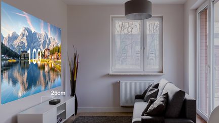 En stue med en grå sofa, et hvidt sofabord, en 100-tommer projekteret skærm, et vindue og en hvid radiator