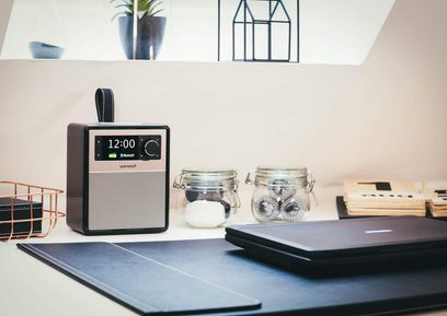 Sonoro Easy sort radio - blank og sølv, set forfra, skærm der viser 12:00, placeret på bord med bærbar computer, glasskrukker, bøger